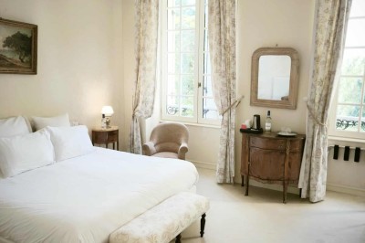 Façade claire avec volets blancs, lanterne en fer forgé et jardinières de fleurs roses en premier plan.