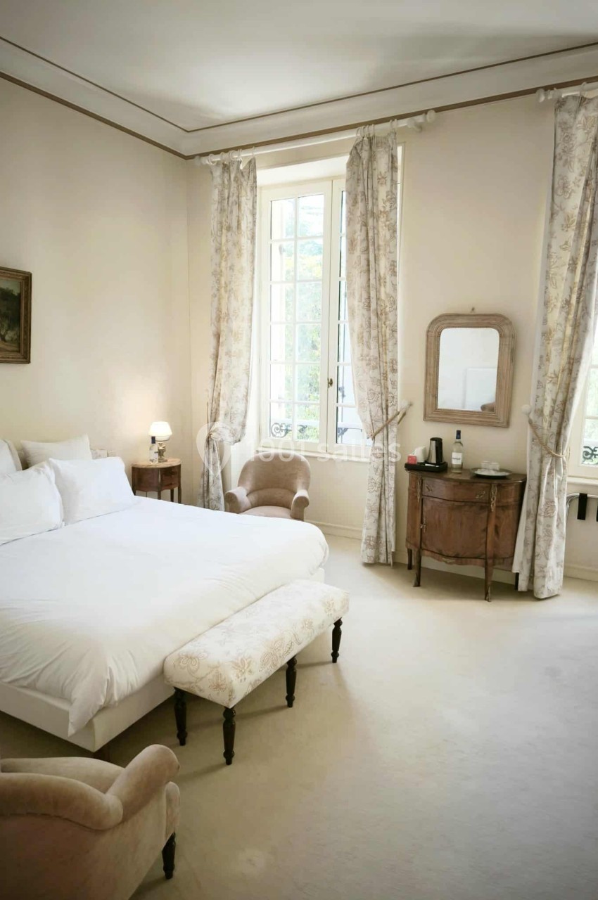 Chambre lumineuse avec lit double, fauteuils, commode, miroir et grandes fenêtres encadrées de rideaux clairs.