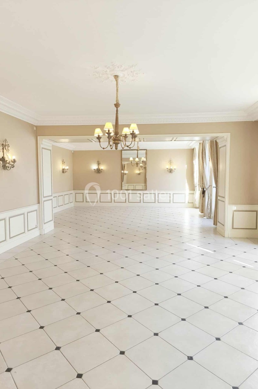 Salle lumineuse avec carrelage clair, moulures au plafond, lustres et grandes fenêtres avec rideaux beiges.