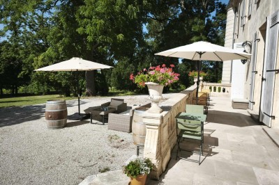 Façade claire avec volets blancs, lanterne en fer forgé et jardinières de fleurs roses en premier plan.
