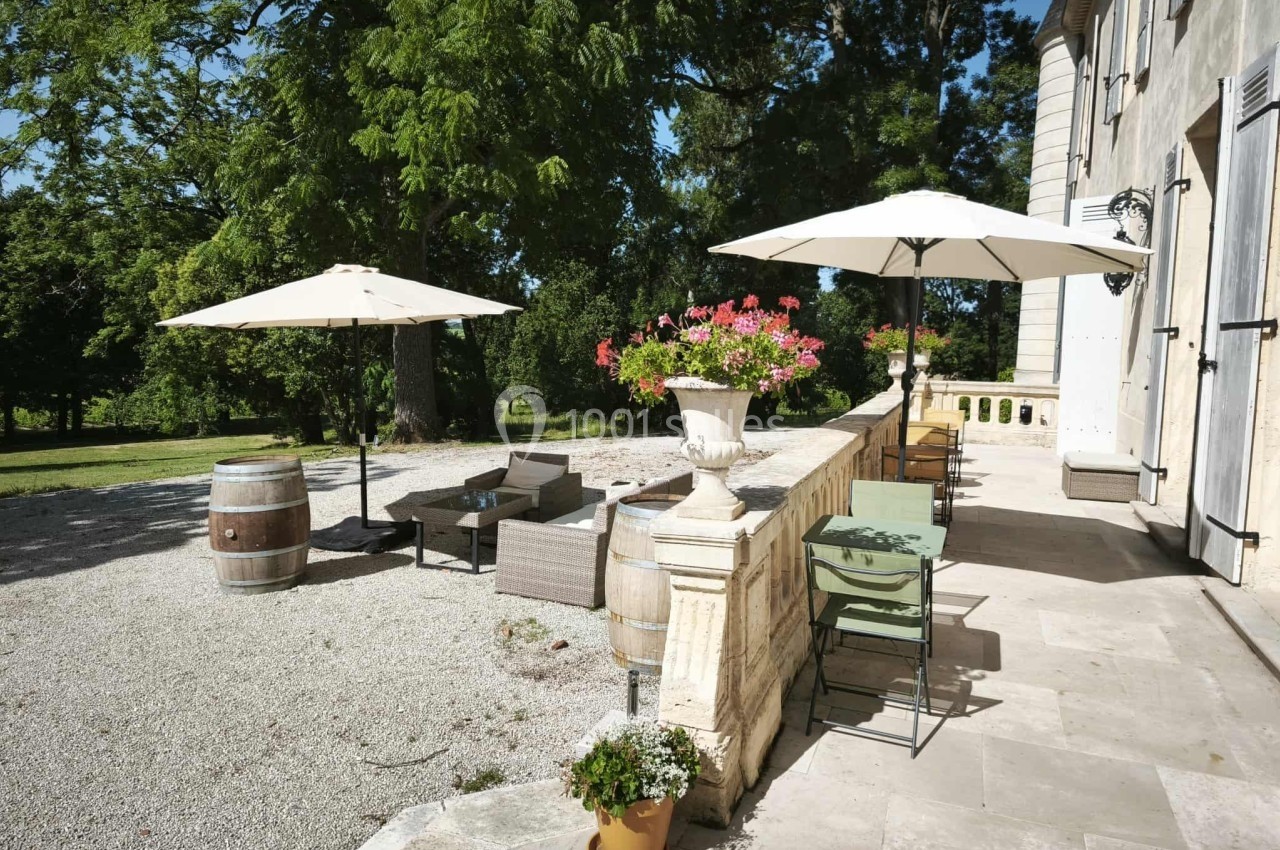 Terrasse ensoleillée avec mobilier de jardin, parasols, pots de fleurs et vue sur un espace vert arboré.