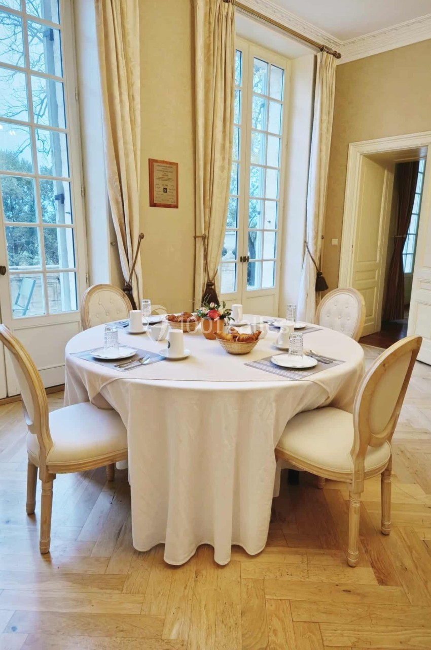 Table ronde dressée avec nappes blanches, chaises élégantes, vaisselle et centre de table floral dans une pièce lumineuse.