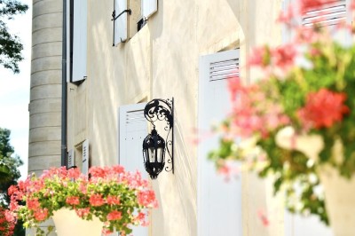Façade claire avec volets blancs, lanterne en fer forgé et jardinières de fleurs roses en premier plan.