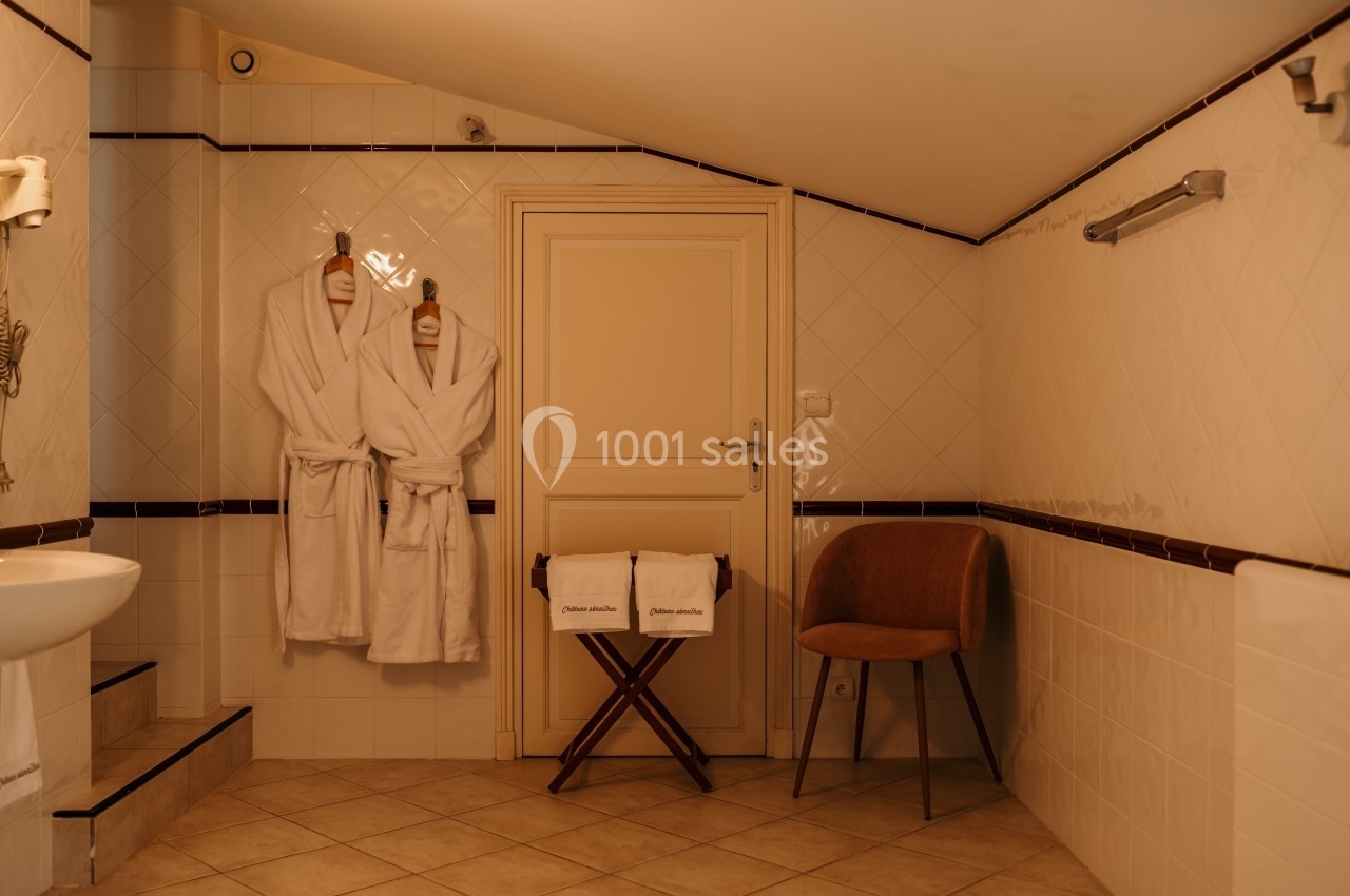 Deux peignoirs accrochés à une porte dans une salle de bain carrelée avec chaise et porte-serviettes.