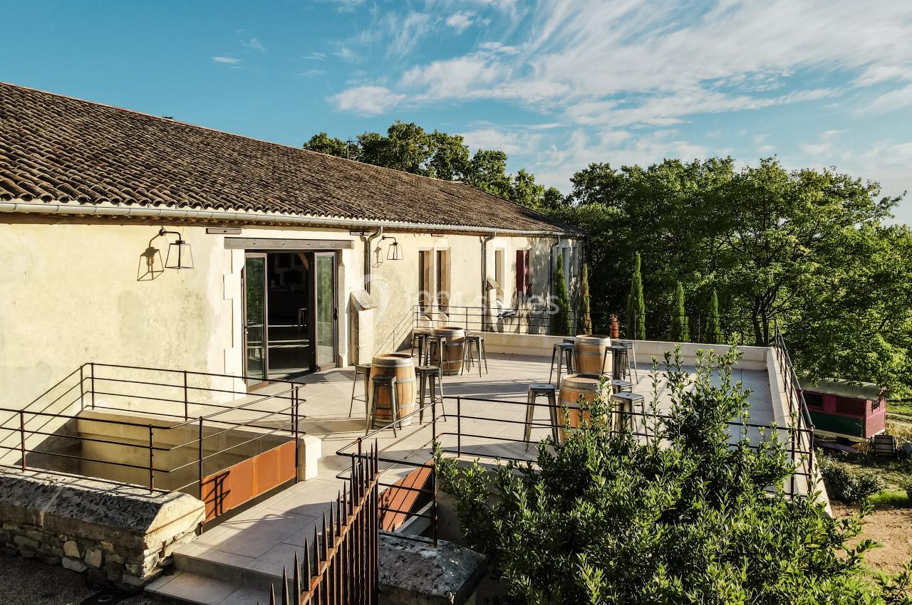 Terrasse ensoleillée avec tables hautes, chaises et vue sur des arbres, attenante à un bâtiment en pierre.