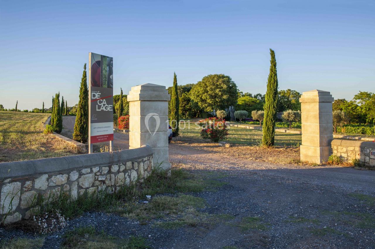 Entrée d'un domaine avec piliers en pierre, allée gravillonnée, cyprès et végétation sous un ciel dégagé.