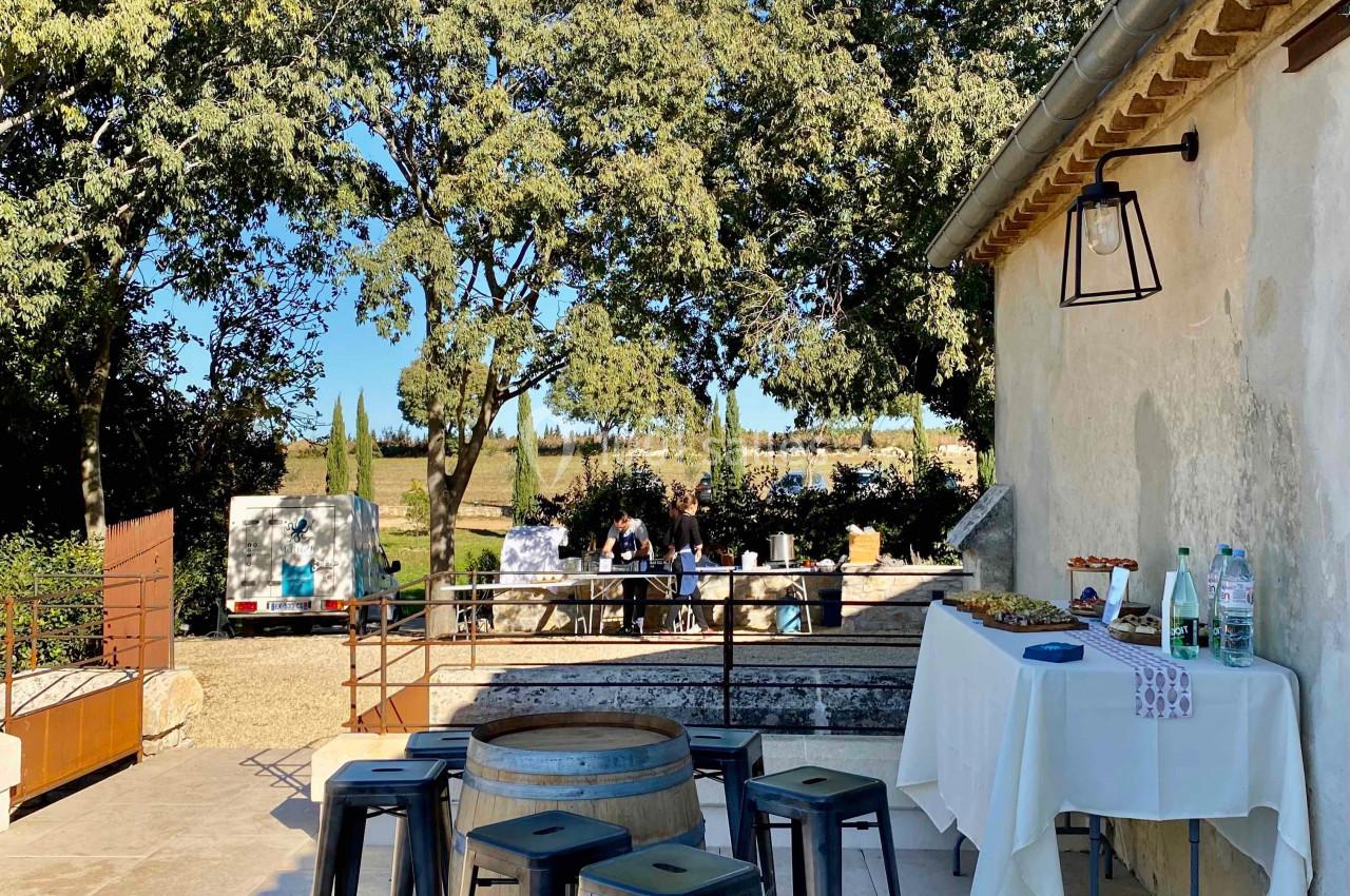 Terrasse ensoleillée avec tables hautes, tabourets, table dressée et vue sur un jardin arboré en arrière-plan.
