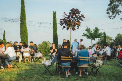 Personnes assises à des tables en plein air dans un jardin, entourées de verdure et de guirlandes lumineuses suspendues.