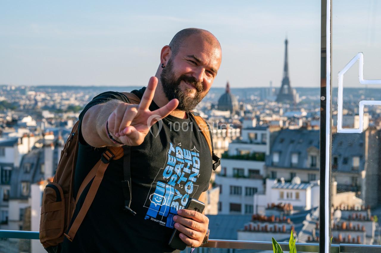 Un homme souriant fait un signe de paix devant une vue panoramique de Paris avec la tour Eiffel en arrière-plan.