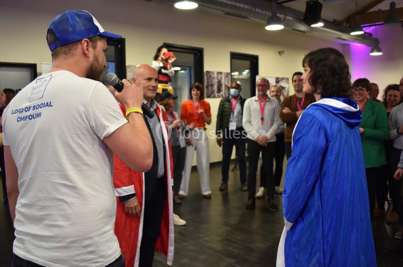 Un homme au micro s'adresse à deux personnes déguisées, entourées d'un groupe de spectateurs dans une salle.