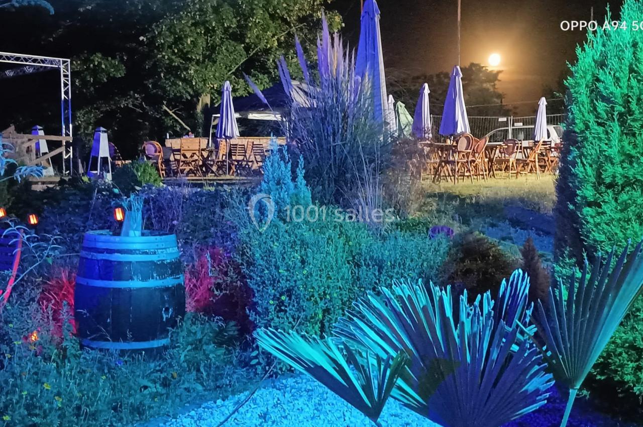 Jardin nocturne illuminé avec végétation, tonneau décoratif et tables en arrière-plan sous une pleine lune.