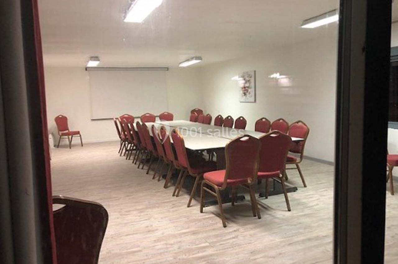 Salle de réunion vide avec une grande table rectangulaire entourée de chaises rouges, éclairée par des néons au plafond.