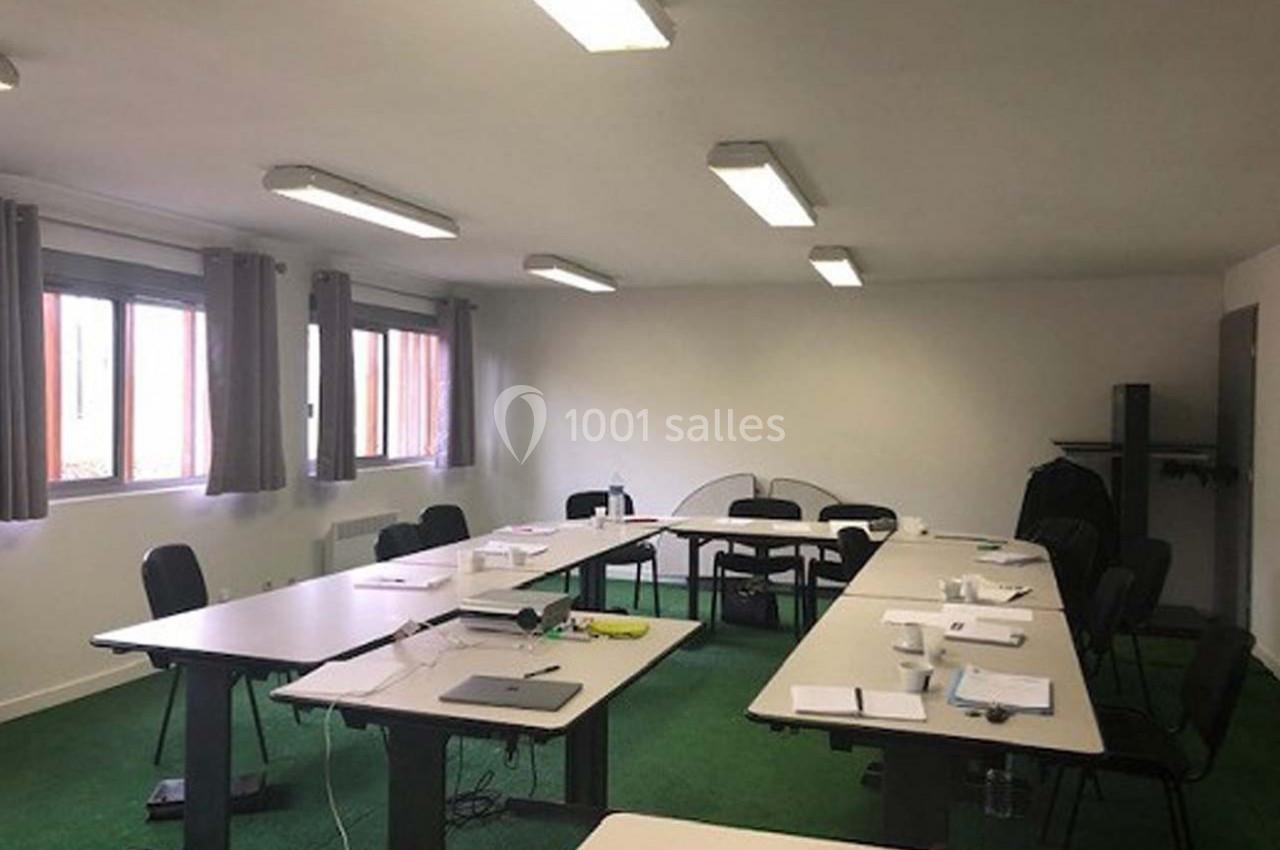Salle de réunion lumineuse avec plusieurs tables disposées en U, chaises noires et fenêtres avec rideaux gris.