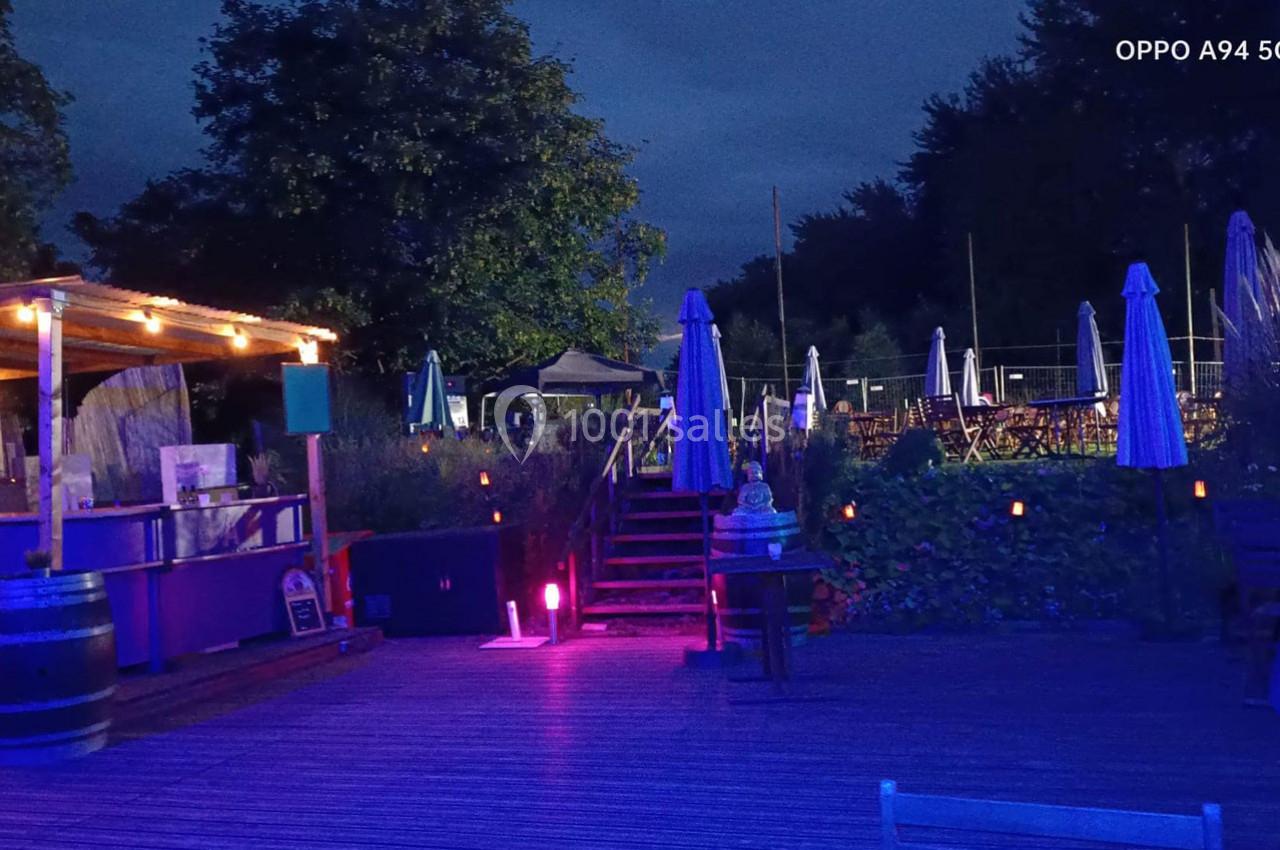 Terrasse extérieure éclairée en soirée avec des parasols, des tables et un bar en bois sous des lumières tamisées.