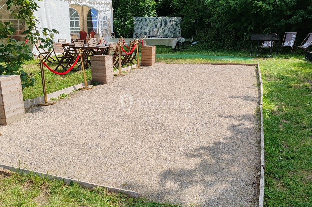 Terrain de pétanque en gravier entouré de verdure, avec des tables et une tente blanche en arrière-plan.