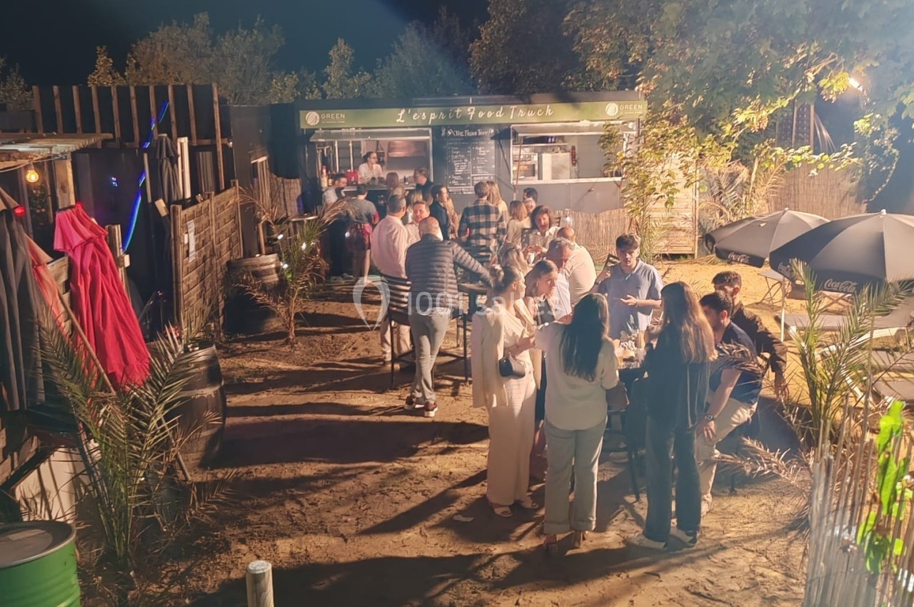 Groupe de personnes rassemblées en soirée devant un food truck, entouré de végétation et éclairé par des lumières.