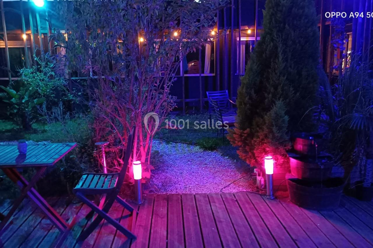 Terrasse en bois éclairée par des lumières colorées, entourée de végétation et de mobilier de jardin.