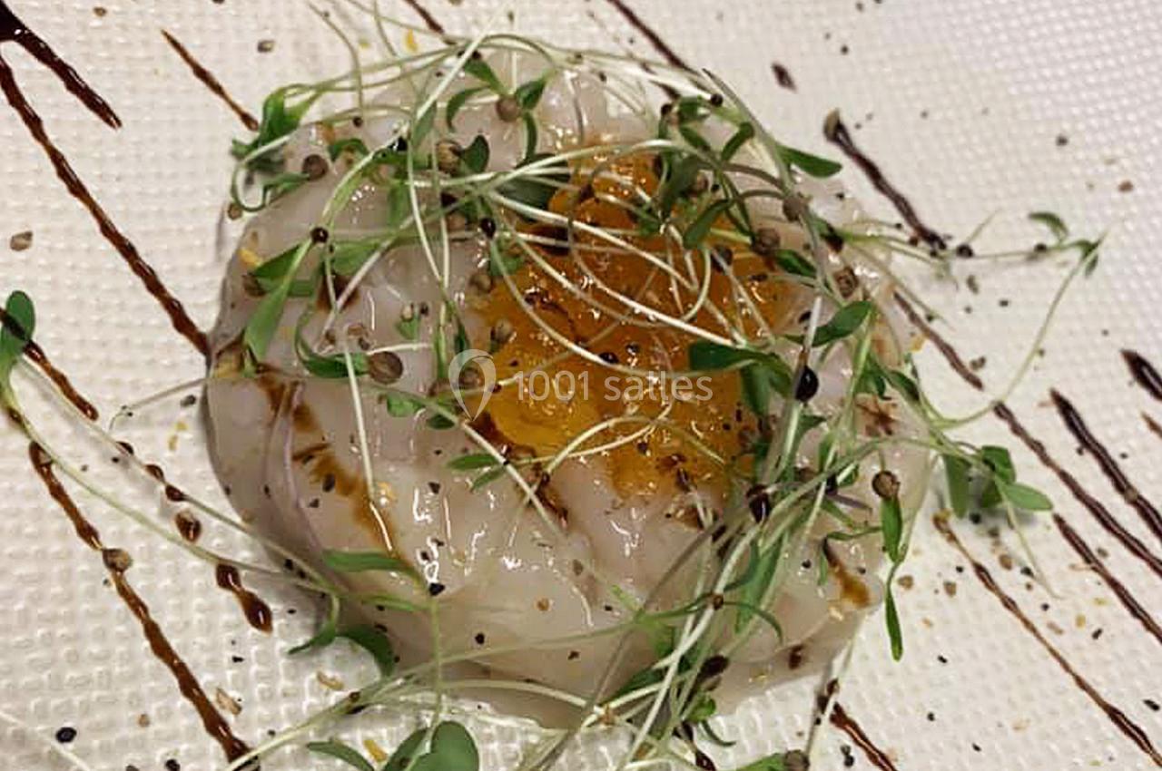 Tranche de carpaccio de Saint-Jacques garnie de pousses vertes, graines et filet de sauce sur assiette texturée.
