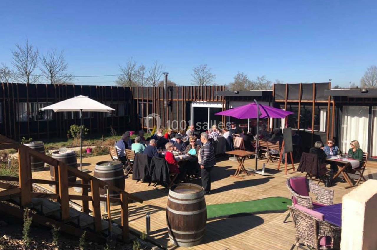 Groupe de personnes assises à des tables en terrasse ensoleillée, entourée de parasols et de végétation.