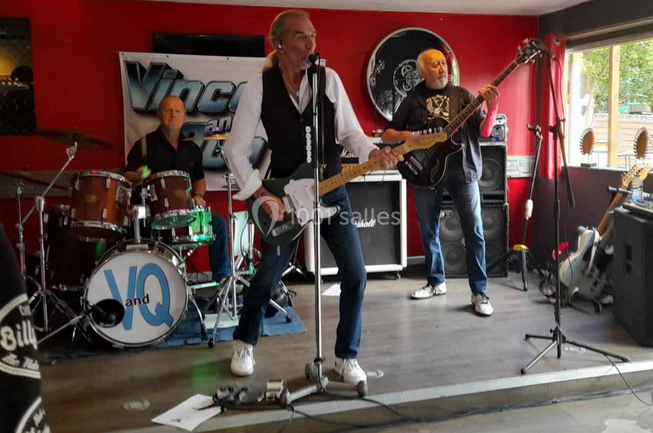 Un groupe de musiciens joue sur une petite scène dans un bar, avec batterie, guitare et basse.