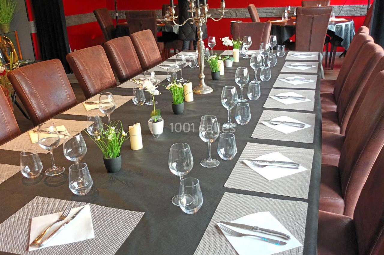 Grande table rectangulaire dressée avec nappes noires, verres, couverts et serviettes, dans une salle de restaurant rouge.