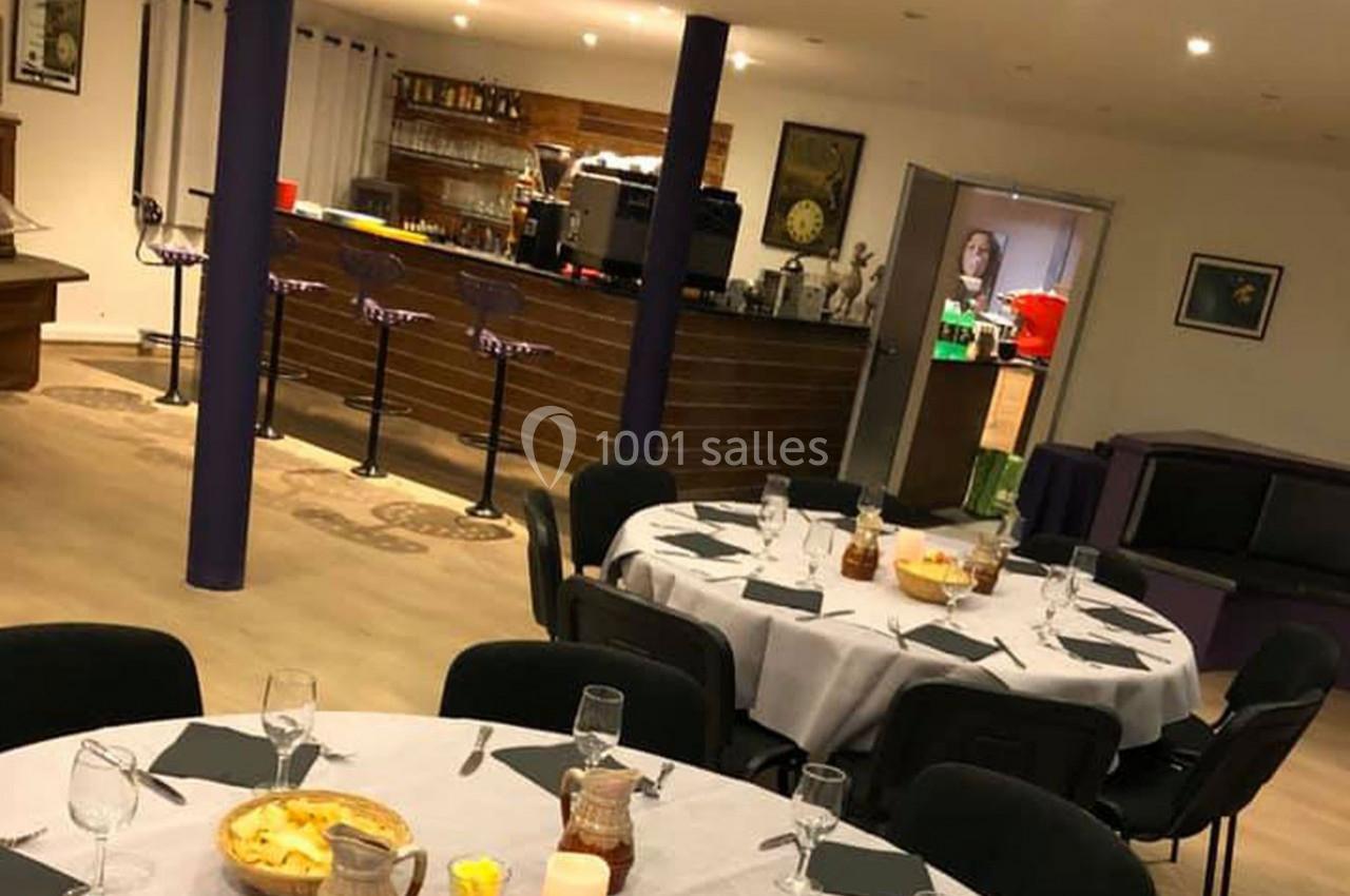 Salle aménagée avec des tables rondes dressées, un bar en arrière-plan et un éclairage chaleureux.