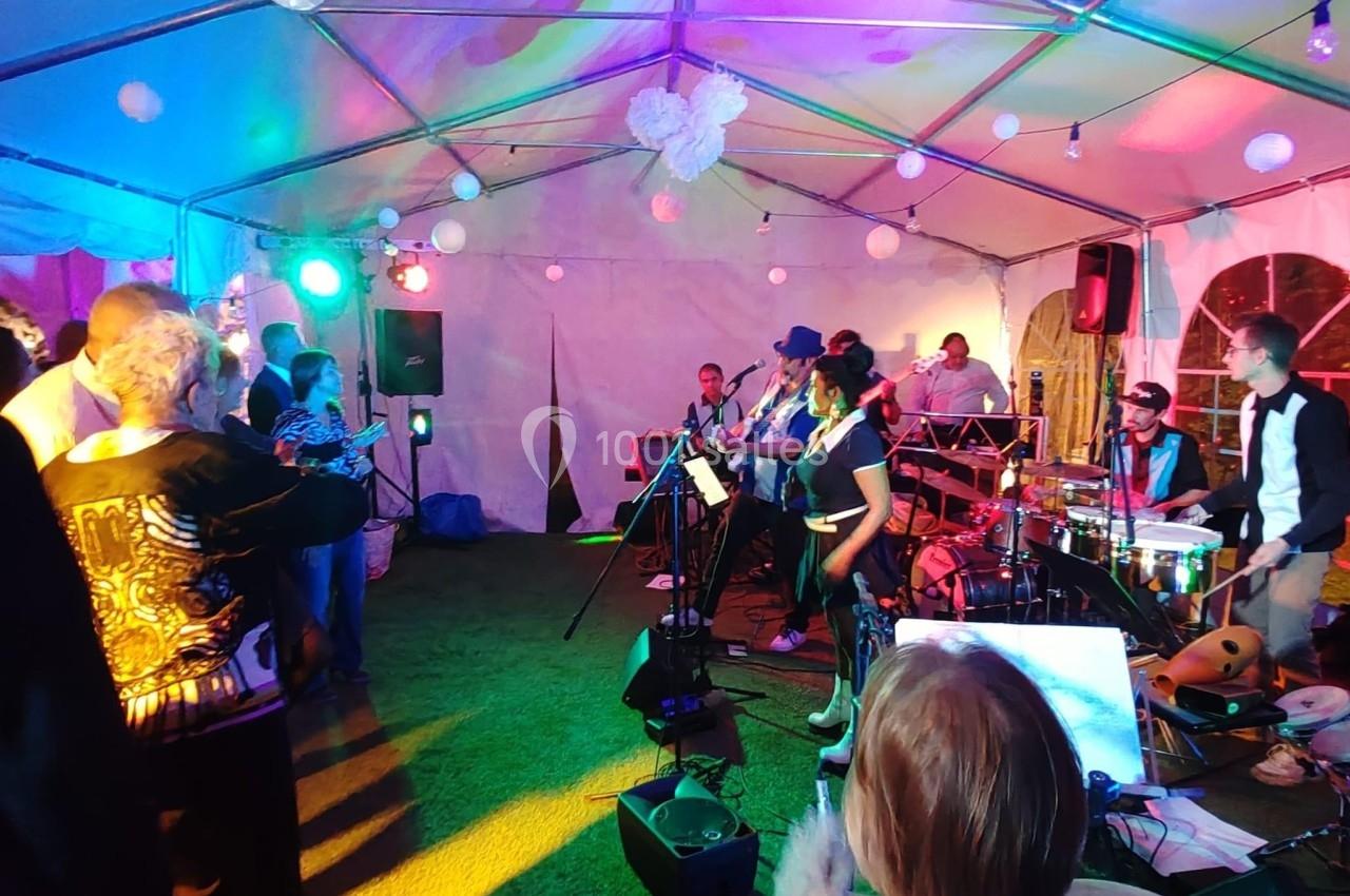 Groupe de musiciens jouant sous une tente éclairée de lumières colorées, avec un public debout les regardant.