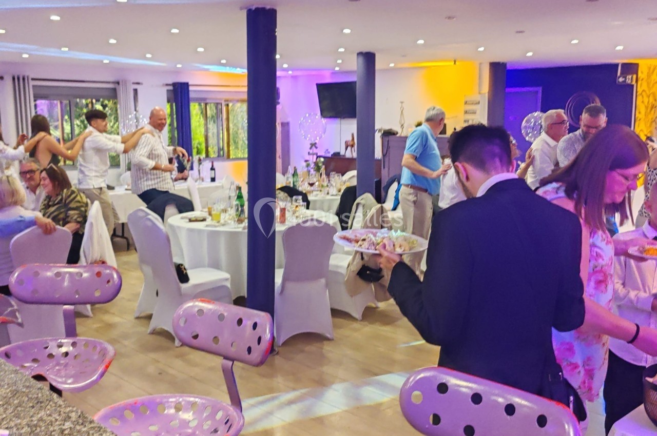 Salle de réception animée avec des invités debout et assis autour de tables décorées, éclairage coloré en arrière-plan.