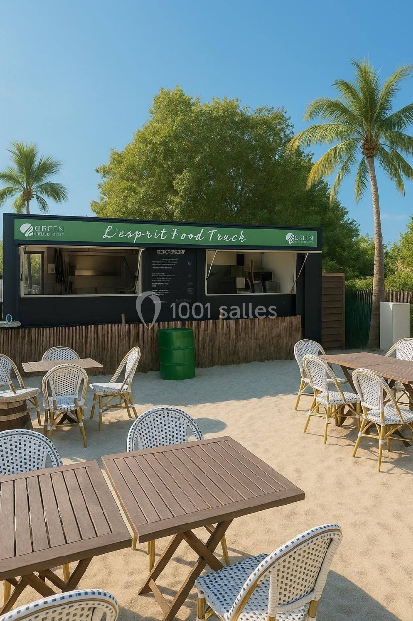 Food truck sur une plage avec des tables et chaises en rotin, entouré de palmiers et d'arbres sous un ciel dégagé.