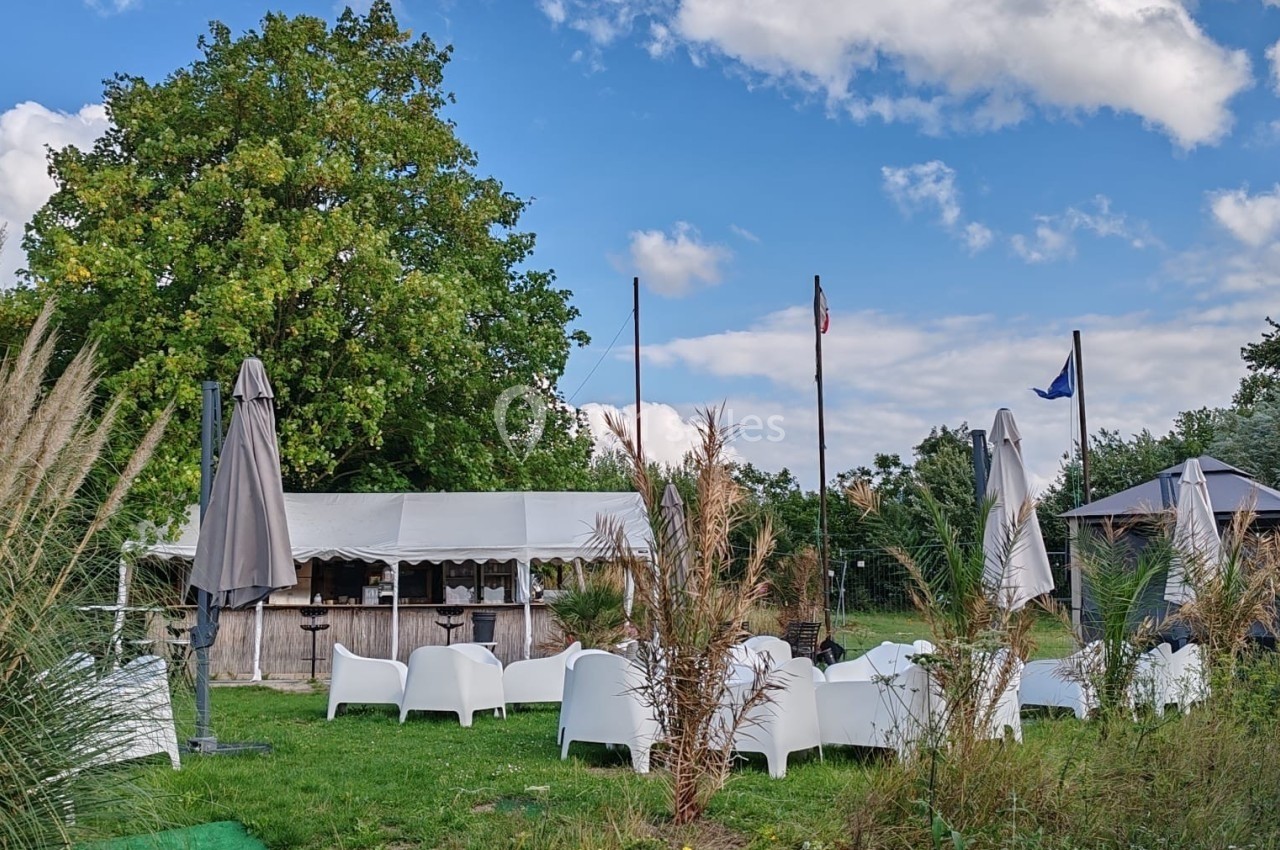 Espace extérieur avec chaises blanches, parasols fermés, tente et drapeaux, entouré de végétation sous un ciel bleu.