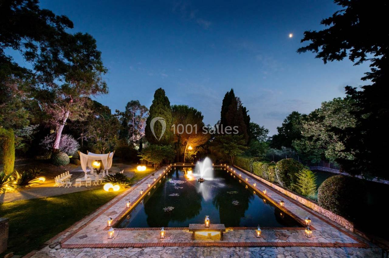 Bassin rectangulaire éclairé par des lanternes, entouré d'arbres, avec une fontaine centrale sous un ciel nocturne.
