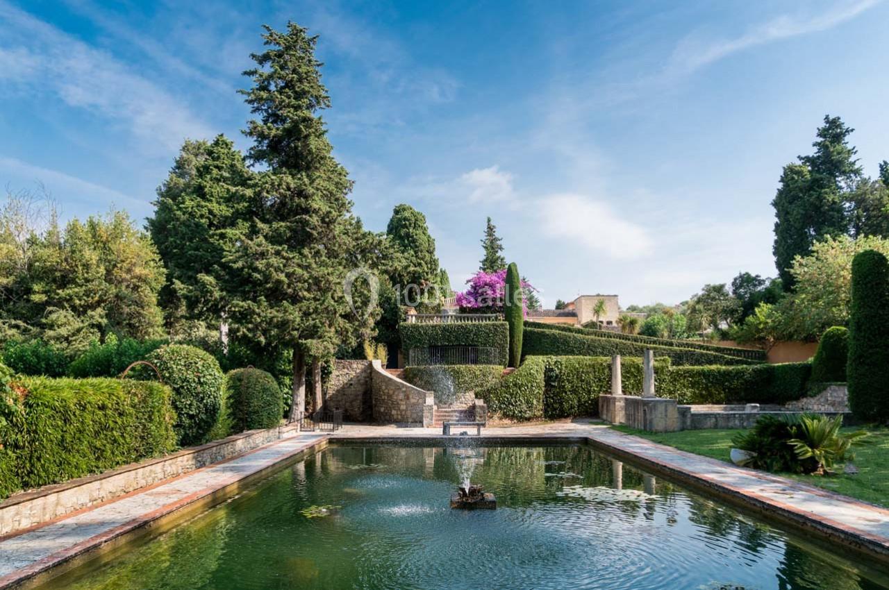 Jardin paysager avec bassin central, fontaine, arbres et buissons taillés, sous un ciel dégagé.