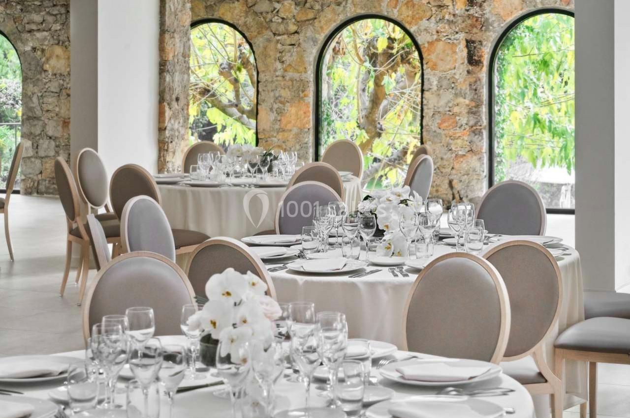 Salle de réception élégante avec tables rondes dressées, nappes blanches, chaises grises et grandes fenêtres en arc.