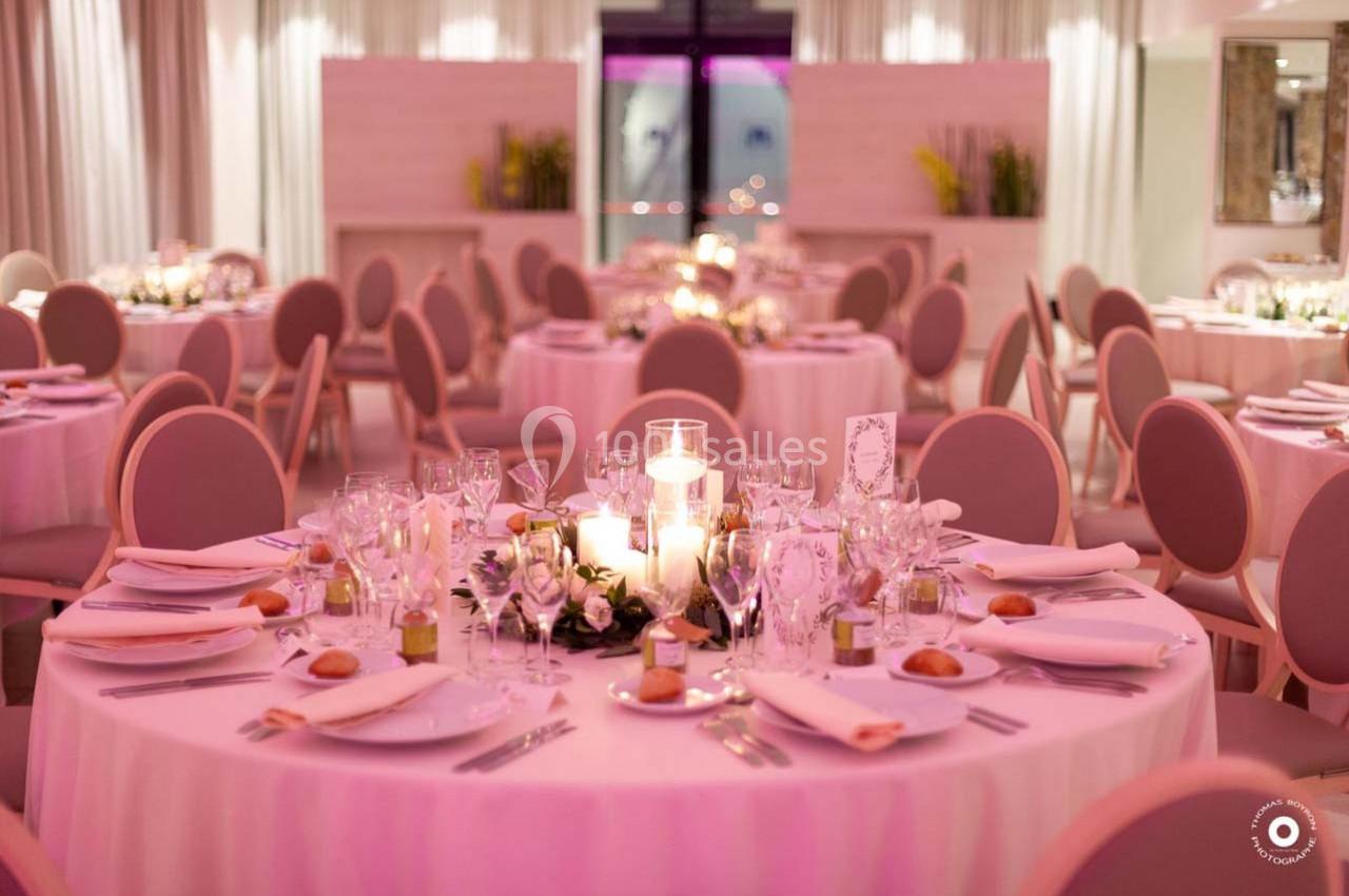 Salle de réception avec tables rondes décorées de bougies, vaisselle élégante et nappes roses.
