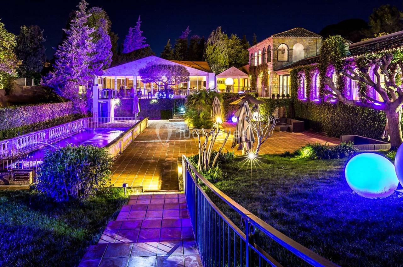 Jardin éclairé de nuit avec piscine, terrasse en pierre et bâtiments entourés de végétation.
