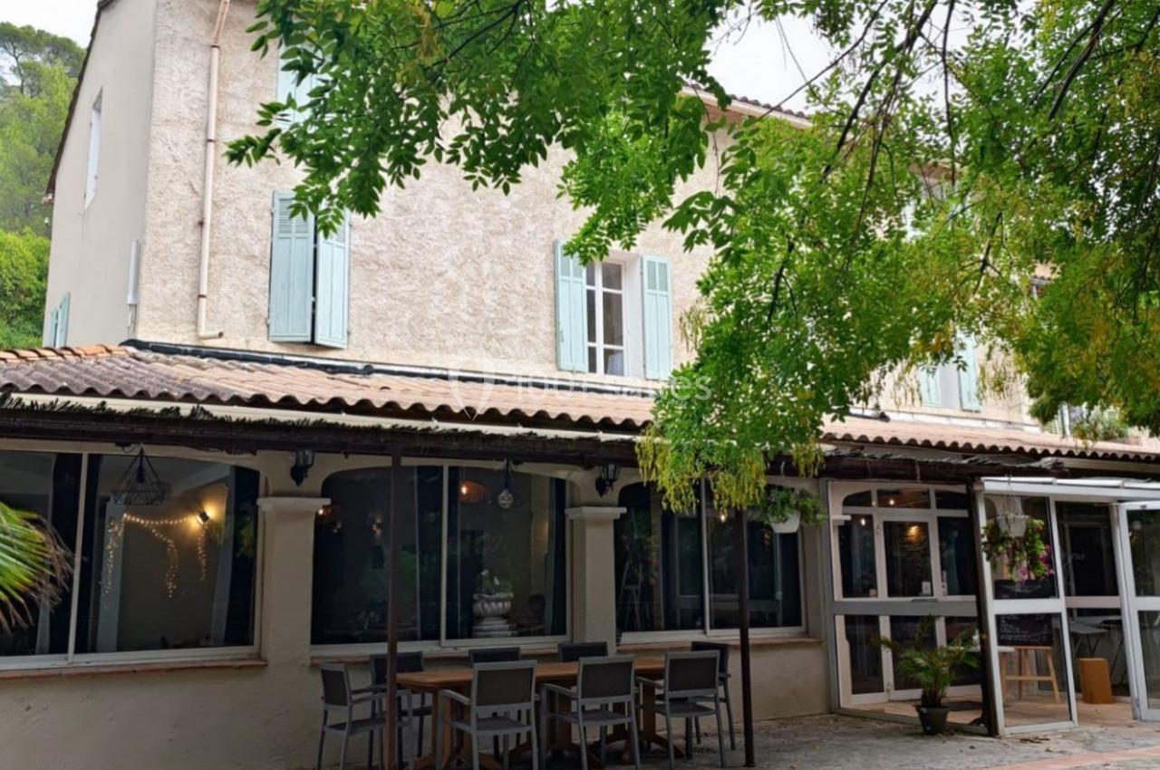 Façade d'une maison provençale avec volets verts, terrasse aménagée et arbres ombrageant l'entrée.