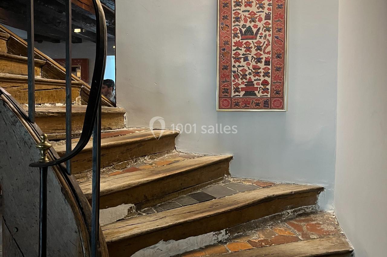 Escalier en bois usé avec une rampe en métal, éclairé par une applique murale, et décoré d'un tapis mural vertical.