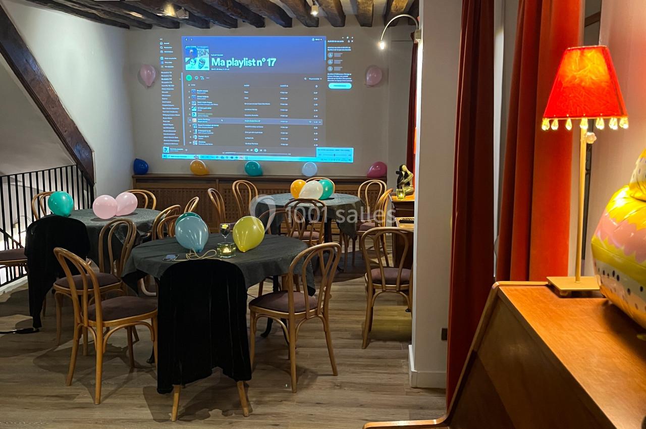 Salle décorée pour une fête avec ballons colorés, tables et chaises en bois, et un écran projetant une playlist musicale.