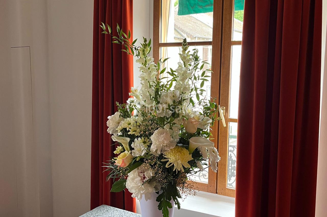 Bouquet de fleurs blanches et jaunes dans un vase blanc, posé sur une table devant une fenêtre encadrée de rideaux rouges.
