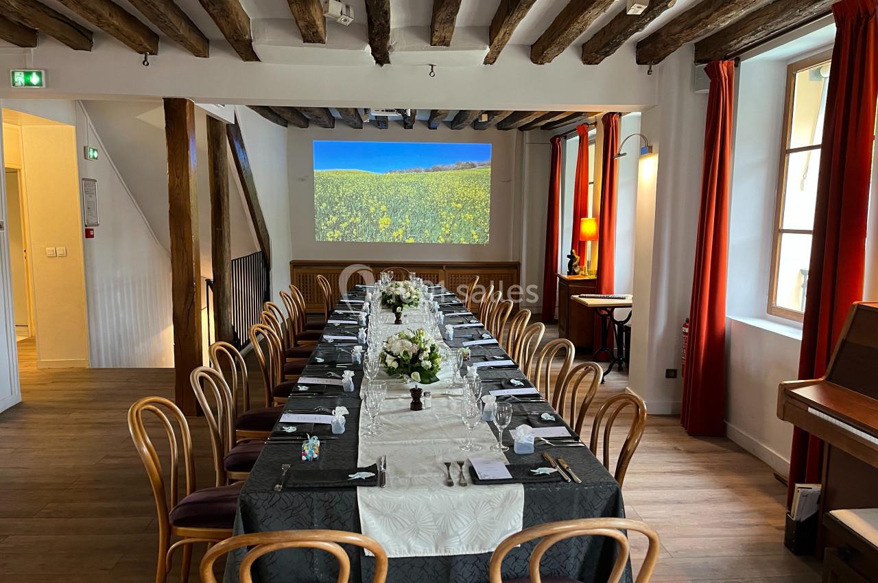 Salle de réception avec une longue table dressée, décorée de fleurs blanches, sous un plafond en poutres apparentes.