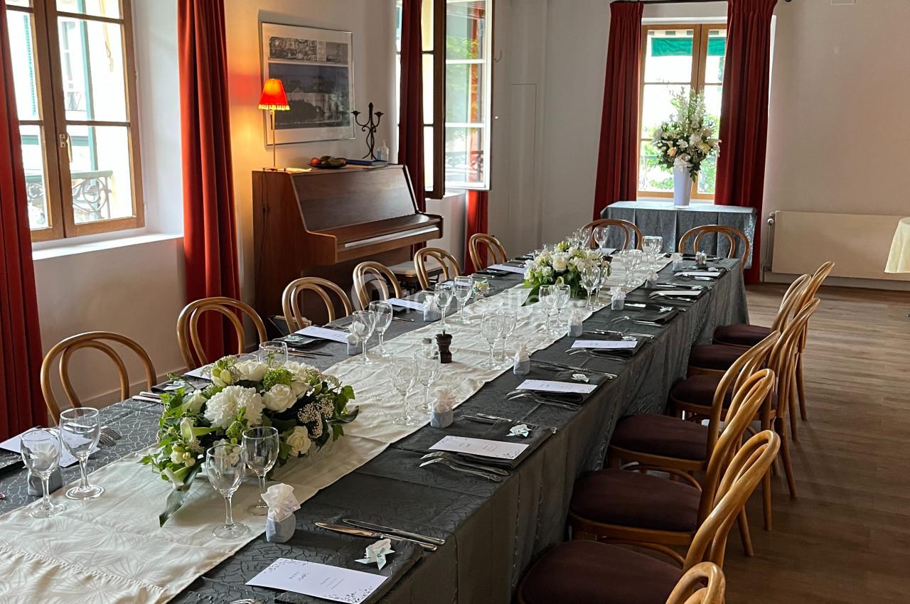 Table dressée dans une salle lumineuse avec poutres apparentes, nappes élégantes, fleurs et chaises en bois.