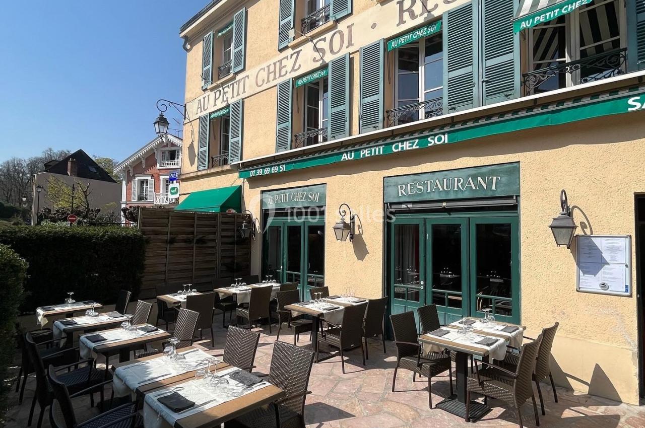 Terrasse ensoleillée d'un restaurant avec des tables dressées, située devant une façade jaune à volets verts.