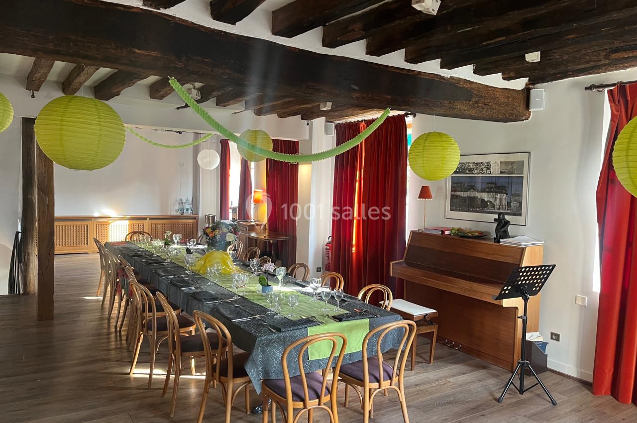 Salle décorée pour un repas festif avec une longue table dressée, des lanternes suspendues et un piano en arrière-plan.