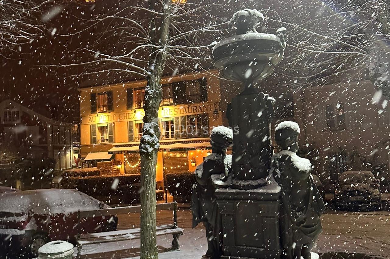 Statue enneigée dans une place éclairée, avec des bâtiments et des décorations lumineuses en arrière-plan.