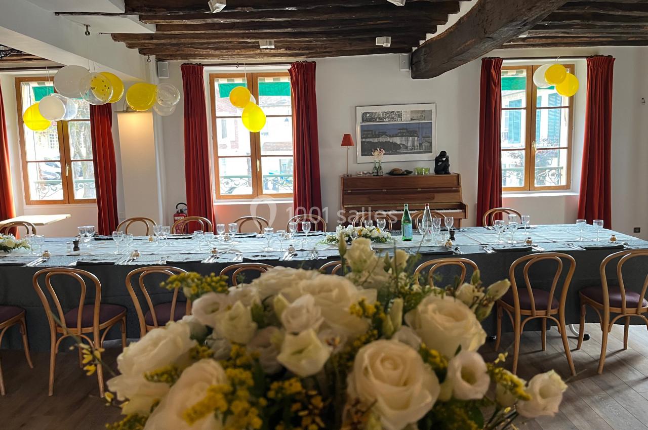 Salle décorée pour un événement avec une grande table dressée, des ballons jaunes et blancs, et un bouquet de fleurs au…