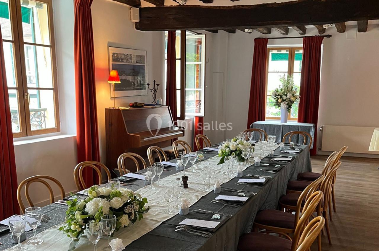 Salle lumineuse avec une grande table dressée pour un repas, décorée de fleurs, entourée de chaises en bois.