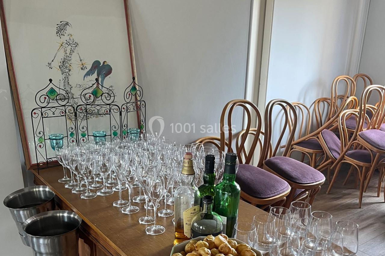 Table avec verres, bouteilles et amuse-bouches, entourée de chaises en bois dans une salle lumineuse.