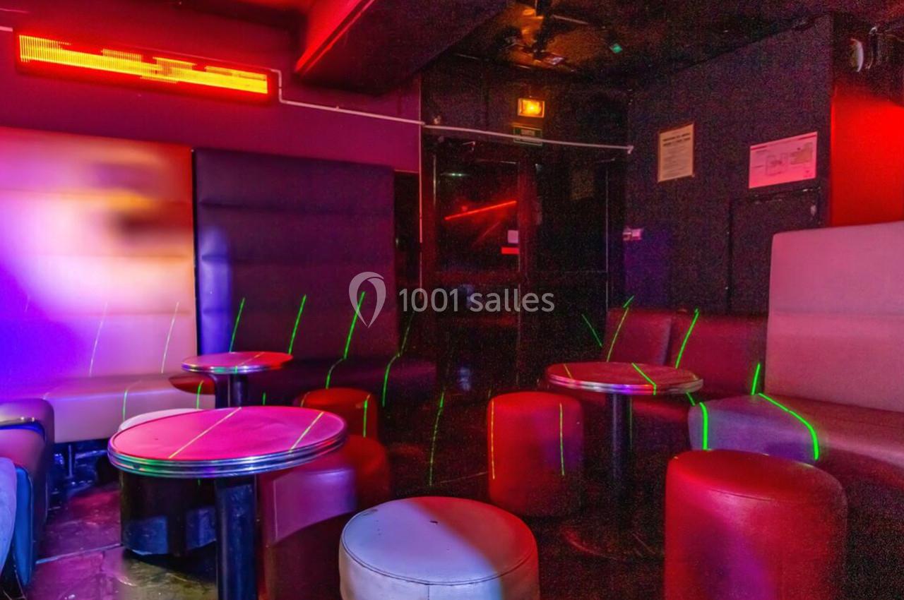 Salle de bar avec banquettes, tables rondes et éclairage tamisé aux tons rouges et violets.