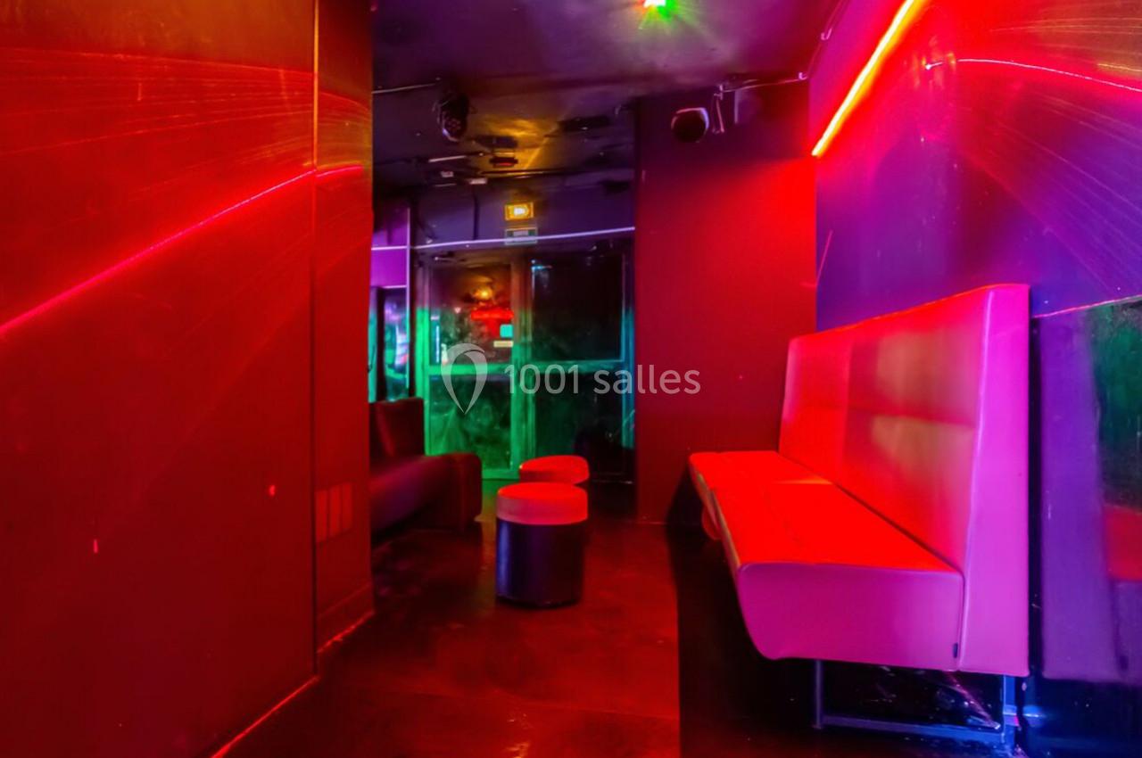 Salle de club avec éclairage néon rouge et violet, banquettes rouges et tabourets noirs sur un sol sombre.