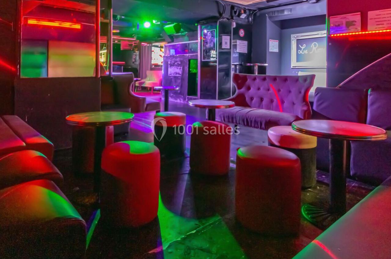 Salle de bar avec canapés, tabourets et tables basses, éclairée par des lumières colorées dans une ambiance tamisée.